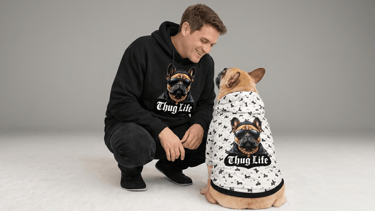 Man smiling with golden retriever and orange cat wearing matching hoodies with playful pet-themed designs.
