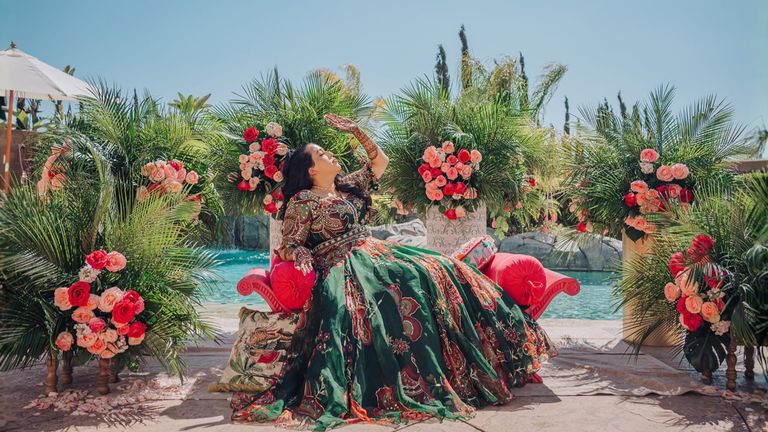 Woman in an emerald gown poses on a floral lounge beside a pool at an elegant outdoor celebration.