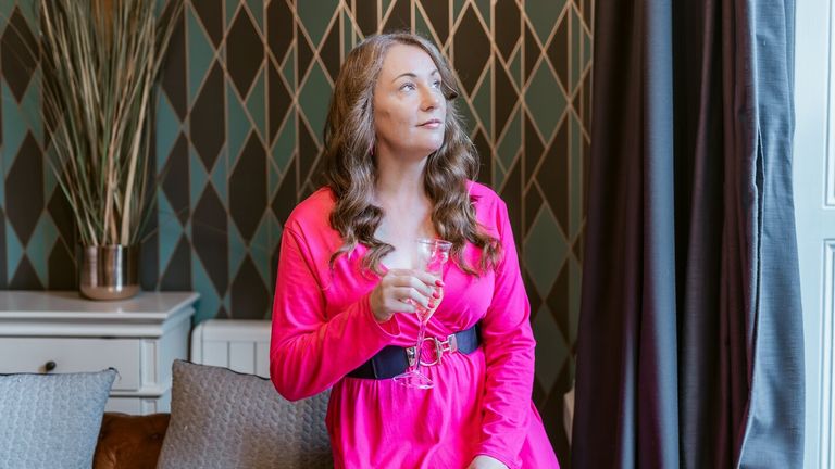 A woman with long wavy hair in a pink dress holding a glass, gazing thoughtfully by a window with a potted plant.