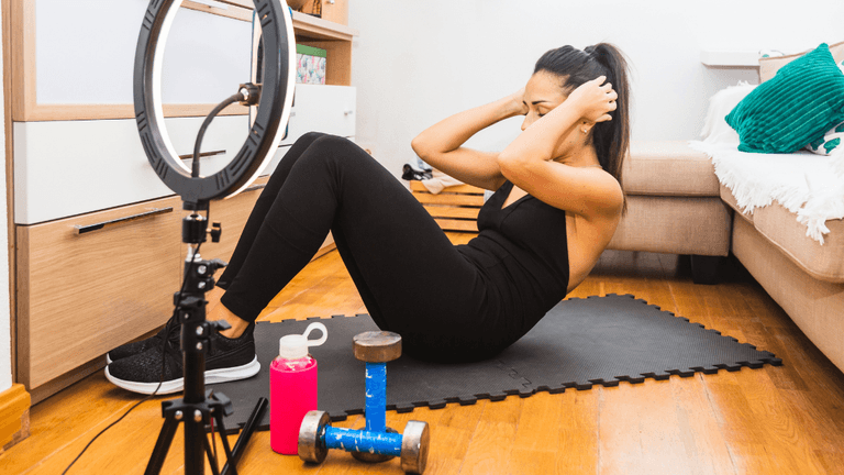 Woman exercising at home while filming a fitness workout with ring light, dumbbells, and sportswear.
