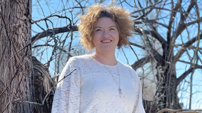 Danielle Dahl standing outside among bare trees, smiling, wearing a white top, symbolizing resilience and healing