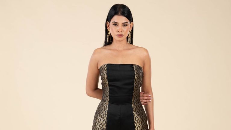 A South Asian woman models a black and gold strapless gown with intricate lace details and bold gold earrings.