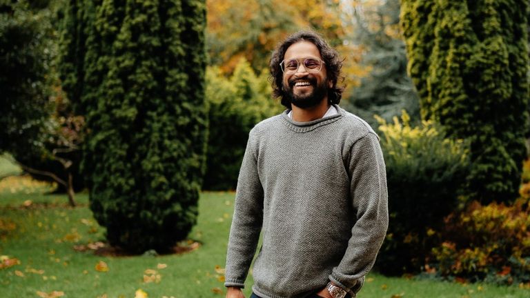A smiling man with glasses stands outdoors amidst green trees, reflecting a warm, approachable presence that connects with nature.