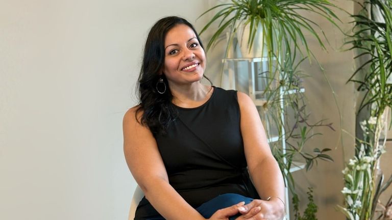 Ellie smiling confidently, dressed in black, seated with plants in the background, embodying her transformational coaching journey.
