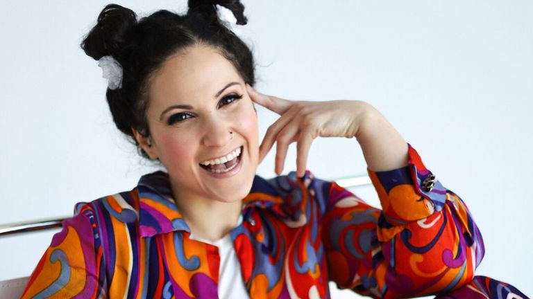 Smiling woman with dark hair in playful buns, wearing a colorful, vibrant shirt with swirling patterns, poses with hand touching her face. Bright and cheerful mood.