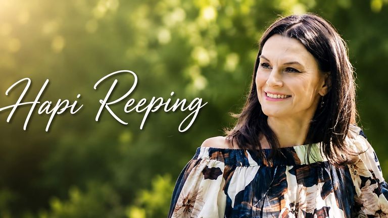 Hapi Reeping smiling outdoors in a floral off-shoulder dress, with “Hapi Reeping” text over a blurred green background.