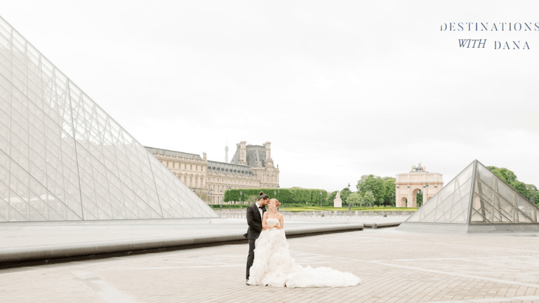 A couple in wedding attire embraces in front of the Louvre Pyramid in Paris, with the Eiffel Tower in the background.