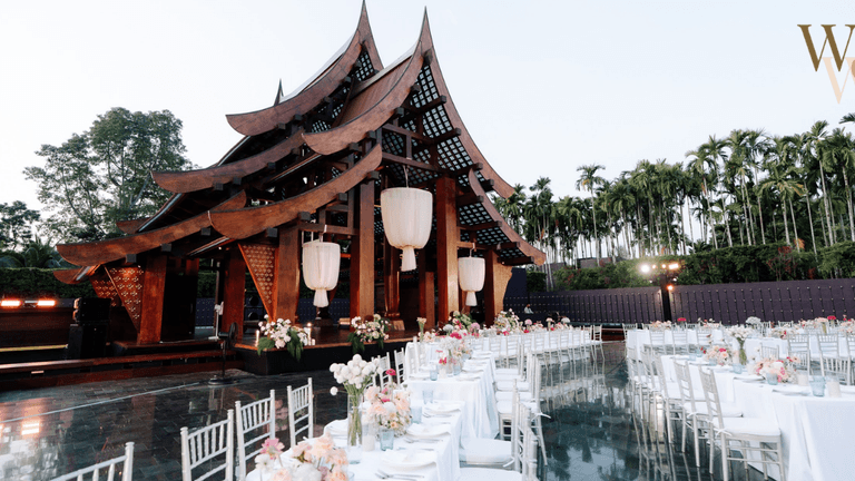 Outdoor wedding venue with a traditional wooden pavilion, long dining tables adorned with floral arrangements, and a scenic backdrop of palm trees under a clear sky.