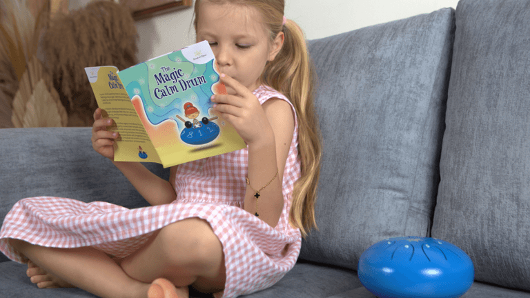 Young girl sitting on a gray couch, reading 'The Magic Calm Drum' book. Next to her is a blue handpan drum. She is dressed in a pink gingham dress and focused on the book, with a calm, relaxed expression.