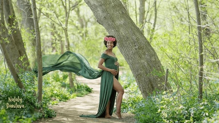 "Pregnant woman in a flowing green dress standing confidently in a nature-filled path with a flower crown, exuding elegance and grace during a maternity photo shoot."