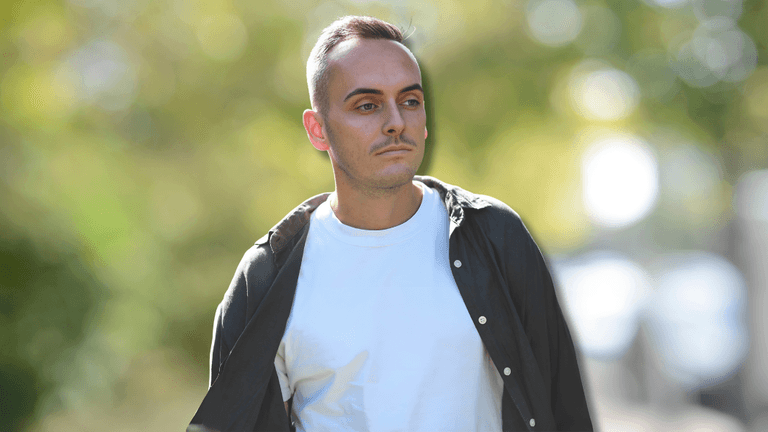 Man walking outdoors on a sunny day with trees and a blurred background.