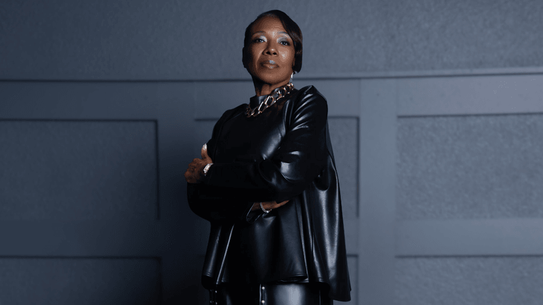Confident woman in a black leather outfit with a gold chain necklace, standing with her arms crossed against a modern, dark gray wall background.