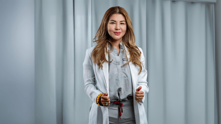 Professional woman in a white lab coat and grey medical scrubs standing confidently with a friendly expression. She is a skilled aesthetician, offering expertise in skincare treatments, with soft lighting and a neutral background.