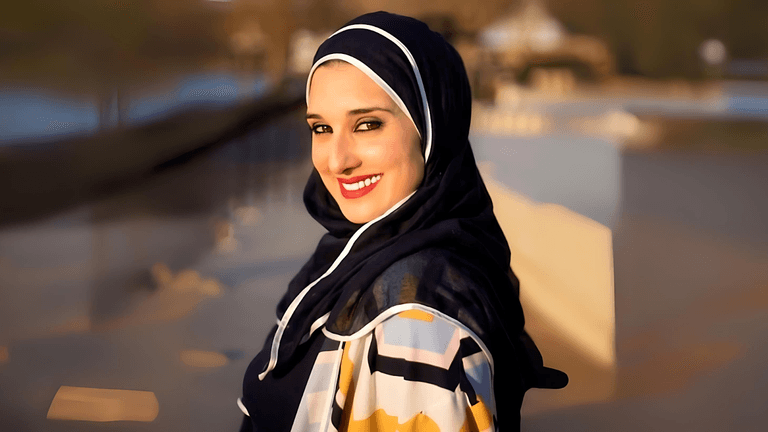 "Smiling woman in a colorful hijab and patterned coat, standing outdoors with a blurred background and sunny lighting."