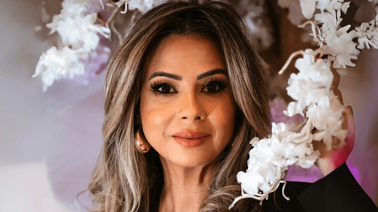 Close-up portrait of a confident woman with glamorous makeup, styled hair, and a warm smile, standing amidst a backdrop of delicate white flowers, radiating elegance and beauty.