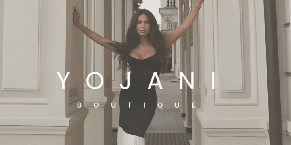 Fashion model posing confidently in a black dress, standing between elegant columns with the 'Yojani Boutique' logo displayed prominently, showcasing sophisticated and stylish clothing.
