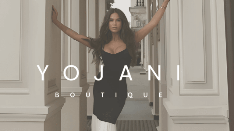 Fashion model posing confidently in a black dress, standing between elegant columns with the 'Yojani Boutique' logo displayed prominently, showcasing sophisticated and stylish clothing.