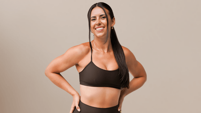 Smiling woman in a dark brown workout outfit, standing with her hands on her hips against a neutral beige background.
