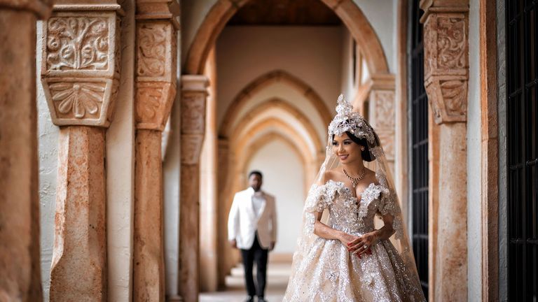 Bride in ornate gown smiles as groom approaches in arched, stone hallway.
