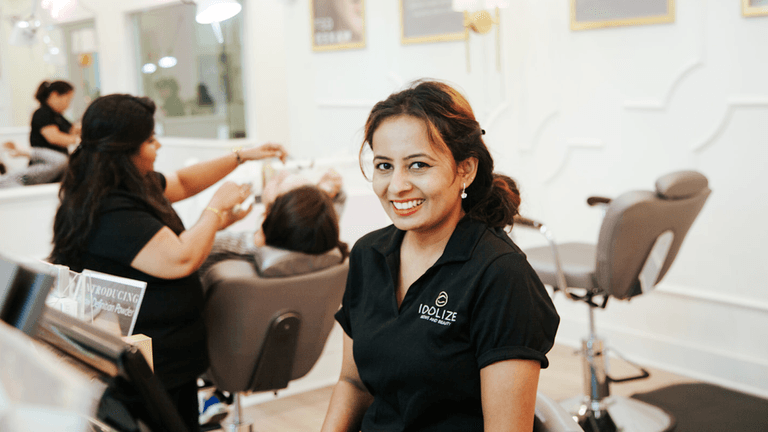 A woman in a black shirt with a logo on it is smiling while sitting at a beauty salon reception desk. In the background, other people are receiving beauty treatments.