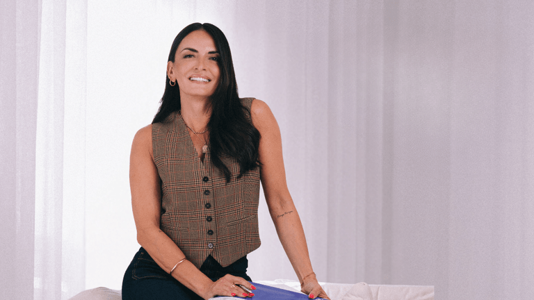 Smiling woman wearing a plaid vest and denim jeans, sitting on a couch with a white background, posing confidently
