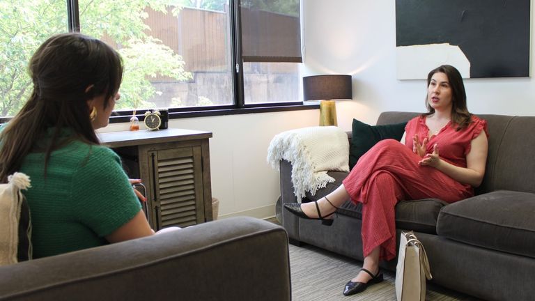 Alt text: "Two women engaged in a conversation in a cozy office setting. One woman, wearing a green sweater, is seated on a gray couch with her back facing the camera. The other woman, dressed in a red striped jumpsuit, is sitting on a separate gray couch, gesturing as she speaks. A wooden cabinet, table lamp, and window with greenery outside are visible in the background.
