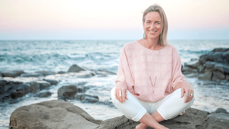 ChatGPT said: Woman in light pink blouse and white pants sitting cross-legged on rocks by the ocean at sunset.