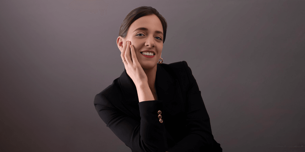 Professional woman in a black suit smiling while gently resting her hand on her face, against a neutral background.