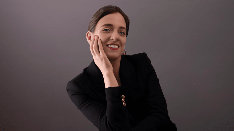 Professional woman in a black suit smiling while gently resting her hand on her face, against a neutral background.