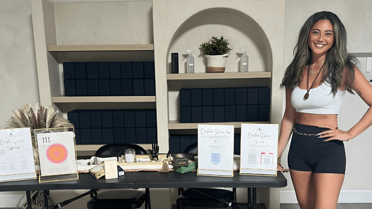 Smiling woman wearing a white crop top and black shorts, standing next to a display table with various crystal products and flyers for her business, with neatly arranged shelves and plants in the background.