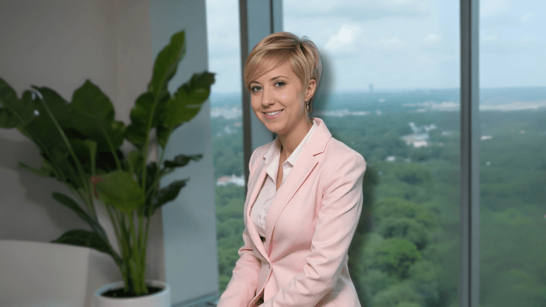 Professional woman with short blonde hair wearing a pink blazer and white blouse, sitting in front of a large window with a city view. She smiles confidently while seated next to a green plant in an office environment.