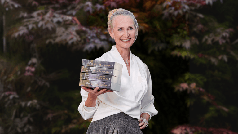 Eva Fellner smiling while holding a stack of The Highlanderess Saga books against a leafy background.