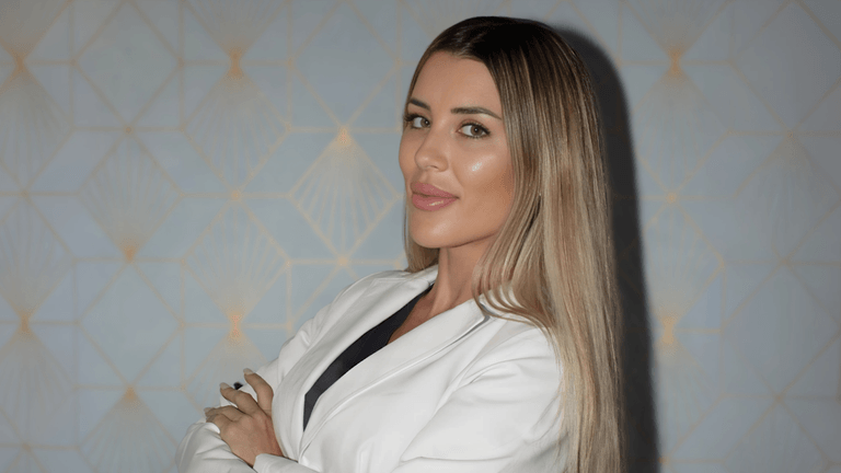 Rita Madozino, founder of Forever Young by Rita, poses confidently in a white coat against a patterned background.