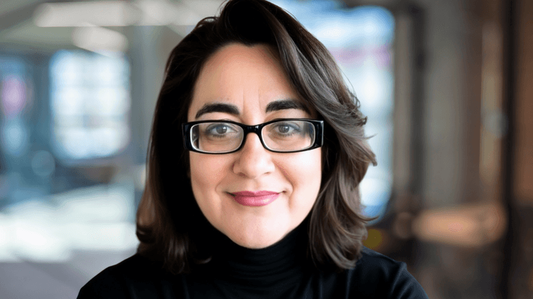Portrait of a woman with brown hair and glasses, wearing a black turtleneck sweater, smiling confidently in a professional setting with blurred background.