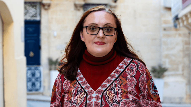Woman dressed in a red turtleneck and patterned blouse, walking through an old city street with a traditional architectural backdrop.