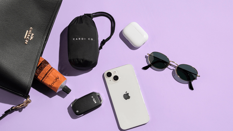 A flat lay of a black Coach handbag, a tube of Aesop hand cream, a "CARDI CO." branded bag, Apple AirPods, a phone, and sunglasses on a purple background.