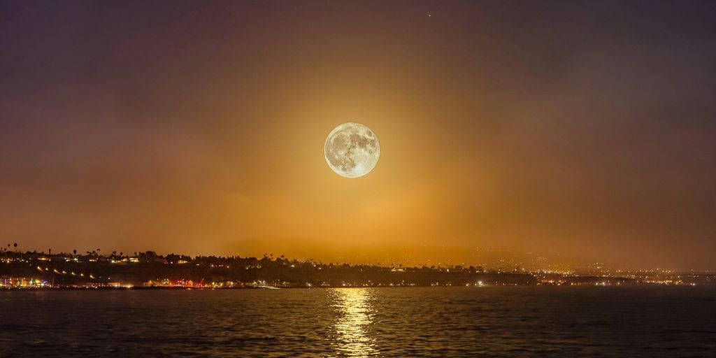 Full moon glowing above a coastal city at night, with golden light reflecting across the water.