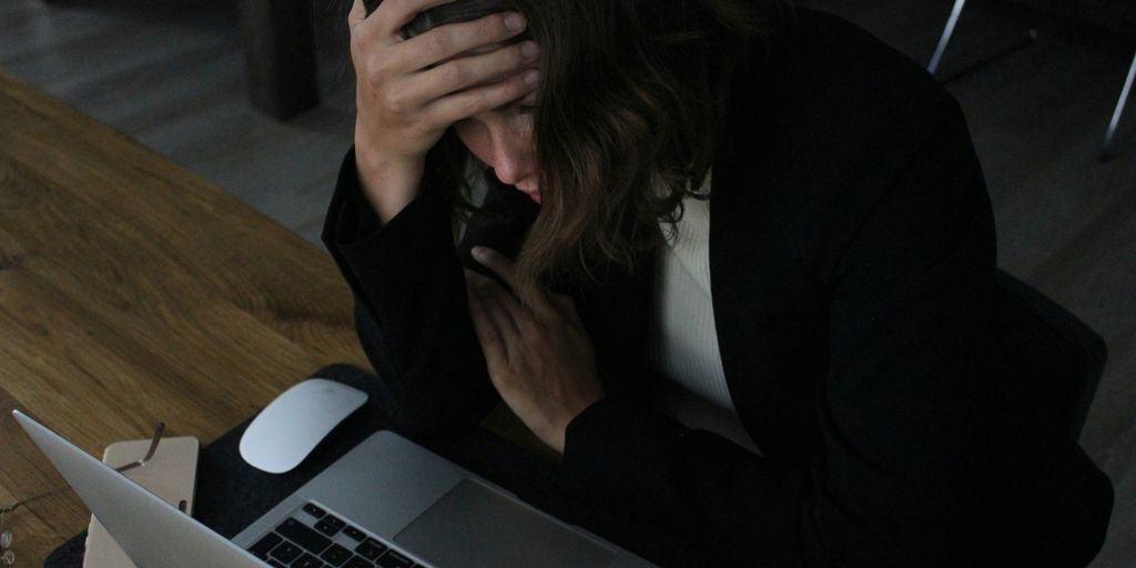 Person sitting at a laptop with one hand on their forehead and the other held against their chest.