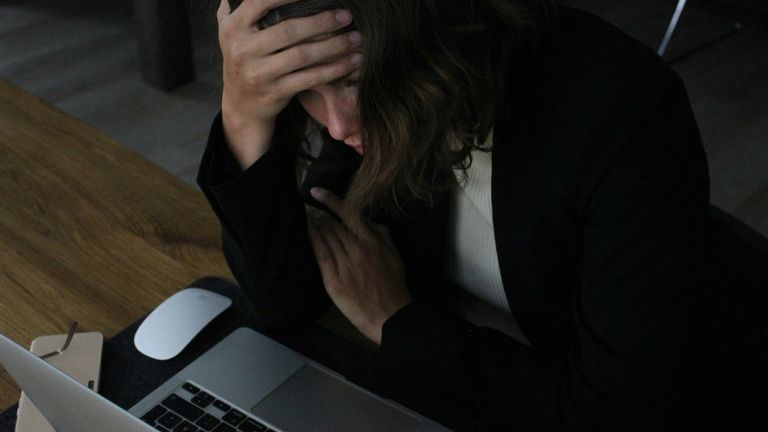 Person sitting at a laptop with one hand on their forehead and the other held against their chest.