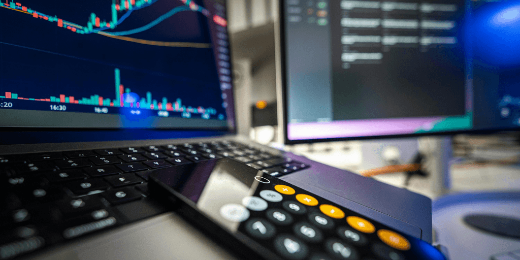 A laptop displaying stock market charts with a calculator and a second monitor.