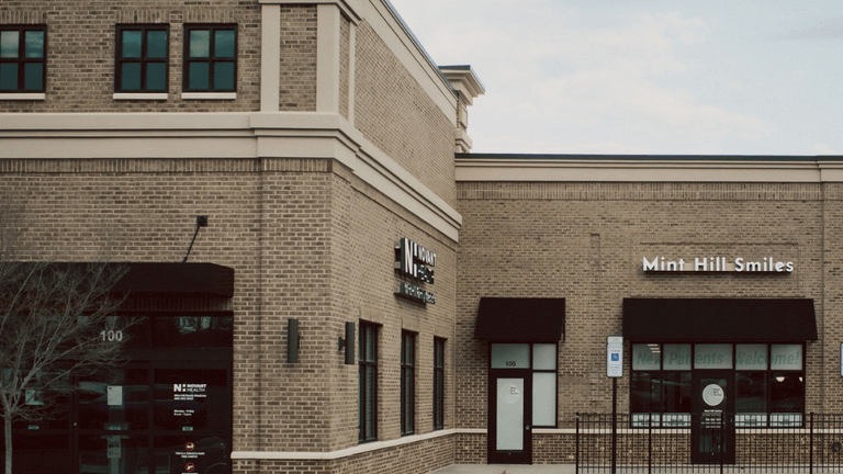 Brick building exterior featuring two storefronts with signs for Mint Hill Smiles and wellness services