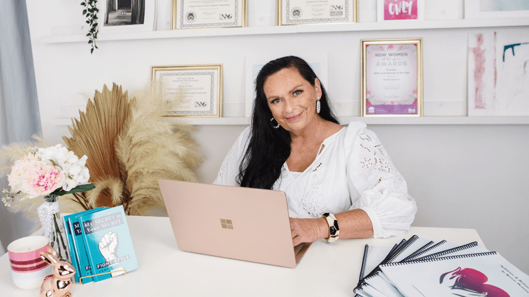 Woman sitting at a desk with awards and notebooks in a bright office setting