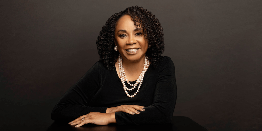 A woman with curly hair, wearing pearls, smiles confidently at the camera.