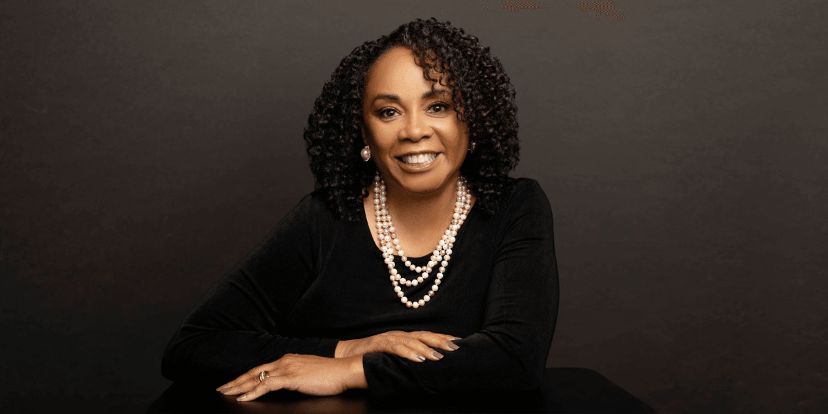 A woman with curly hair, wearing pearls, smiles confidently at the camera.