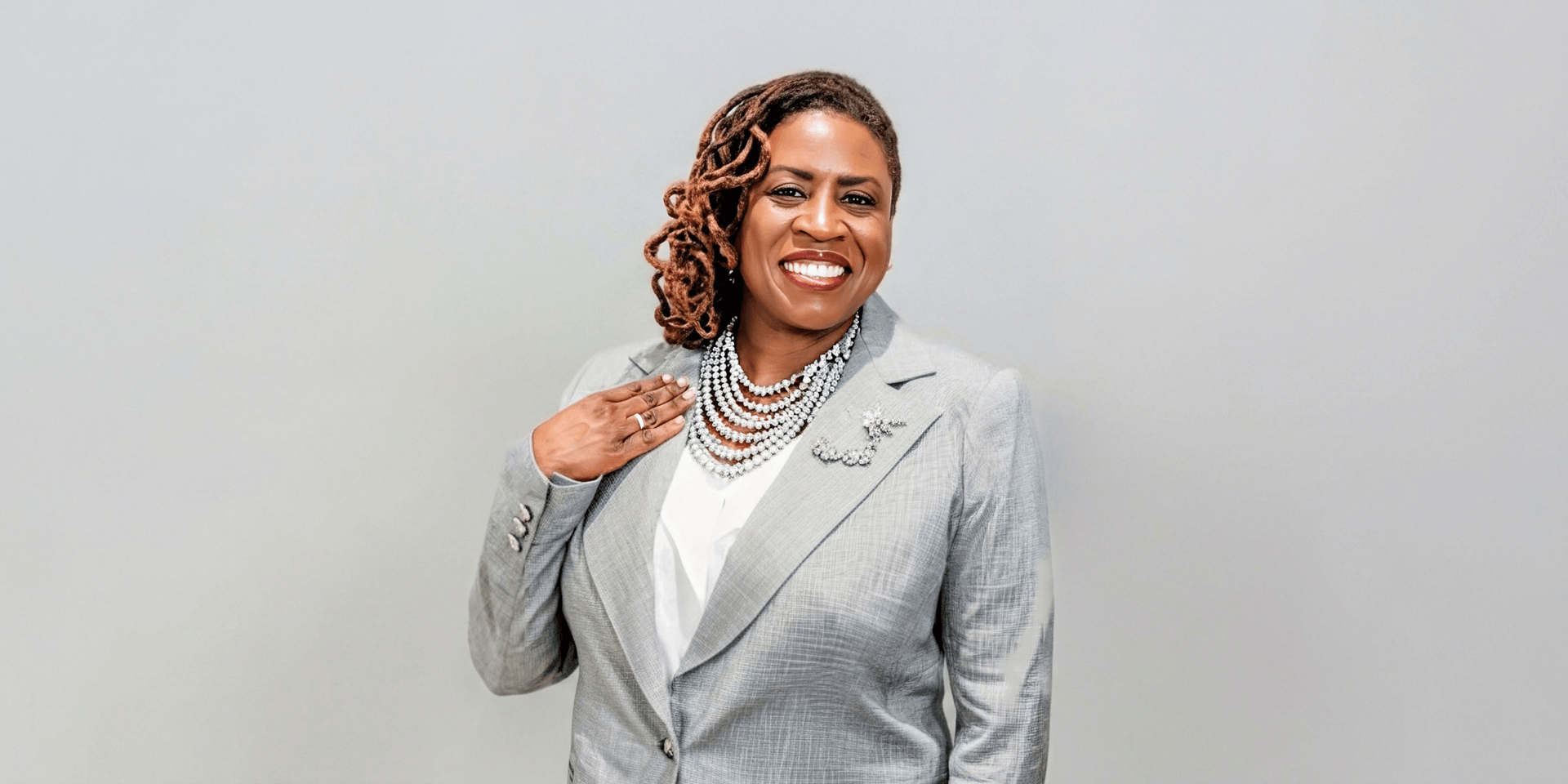 Woman in a gray suit with layered necklaces smiling against a light background