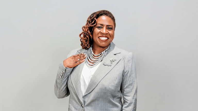 Woman in a gray suit with layered necklaces smiling against a light background