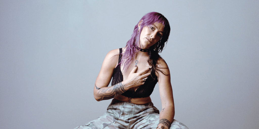 A woman with purple hair and tattoos poses seated against a gray background