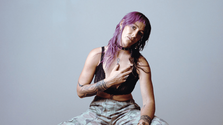 A woman with purple hair and tattoos poses seated against a gray background