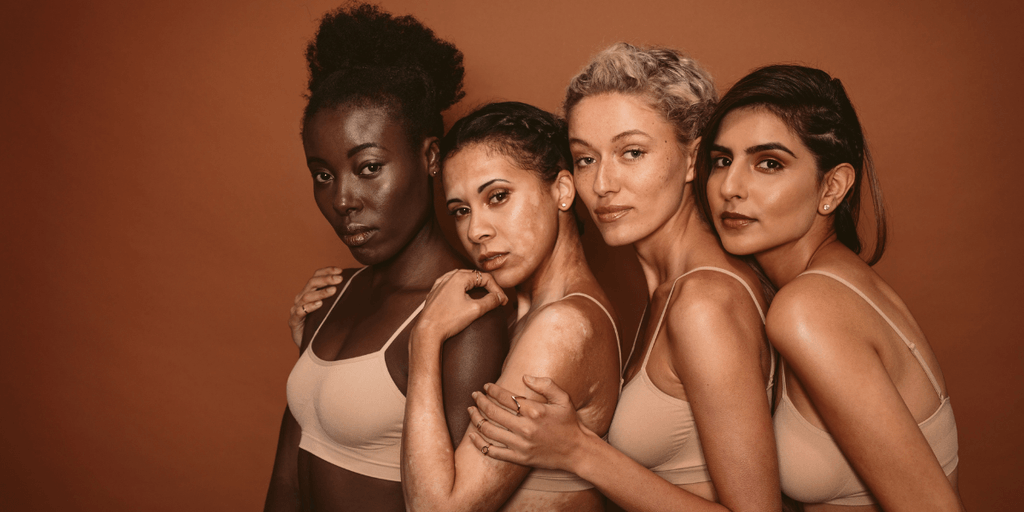 Four women of diverse backgrounds pose together, wearing matching beige tops against a warm backdrop
