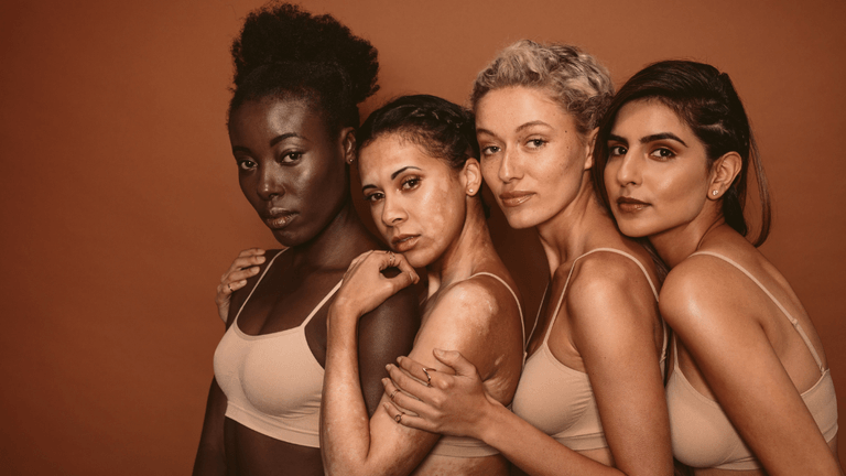 Four women of diverse backgrounds pose together, wearing matching beige tops against a warm backdrop